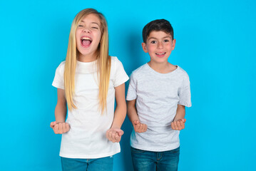 two kids boy and girl standing over blue studio background raising fists up screaming with joy...