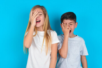 Charismatic carefree joyful two kids boy and girl standing over blue studio background likes laugh out loud not hiding emotions giggling hear funny hilarious joke chuckling facepalm. - obrazy, fototapety, plakaty