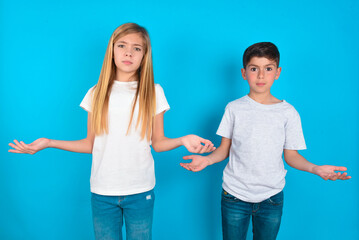 two kids boy and girl standing over blue studio background looks uncertain shrugs shoulders.