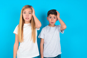 Portrait of confused two kids boy and girl standing over blue studio background holding hand on...