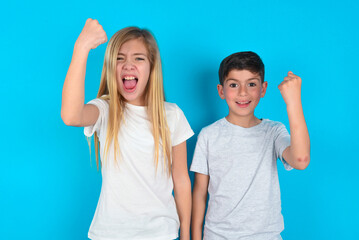 Fototapeta na wymiar Fierce two kids boy and girl standing over blue studio background holding fist in front as if is ready for fight or challenge, screaming and having aggressive expression on face.
