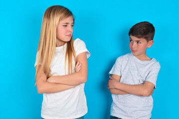 Displeased two kids boy and girl standing over blue studio background with bad attitude, arms crossed looking sideways. Negative human emotion facial expression feelings.