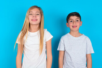 two kids boy and girl standing over blue studio background with happy and funny face smiling and showing tongue.