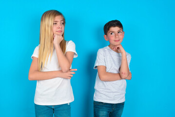 two kids boy and girl standing over blue studio background with hand under chin and looking sideways with doubtful and skeptical expression, suspect and doubt.