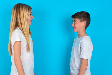 Profile of smiling two kids boy and girl standing over blue studio background with healthy skin, has contemplative expression, ready to have outdoor walk.