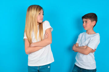 Image of upset two kids boy and girl standing over blue studio background with arms crossed. Looking with disappointed expression aside after listening to bad news.