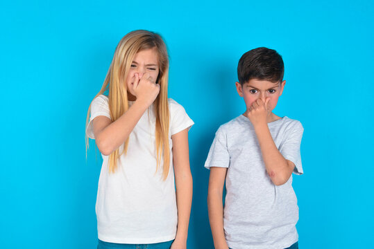 two kids boy and girl standing over blue studio background, holding his nose because of a bad smell.
