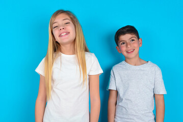 two kids boy and girl standing over blue studio background with broad smile, shows white teeth,...