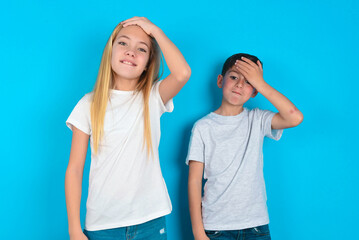 Oops, what did I do? two kids boy and girl standing over blue studio background holding hand on forehead with frightened and regret expression.