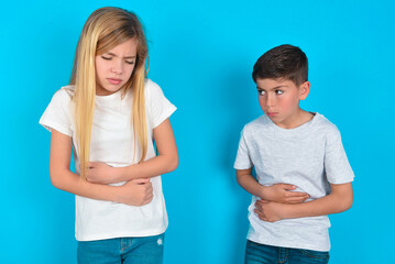 two kids boy and girl standing over blue studio background suffering from strong stomachache.
