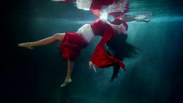 a brunette with long hair and wearing a red suit with white fur floats under the surface of blue water. the general plan