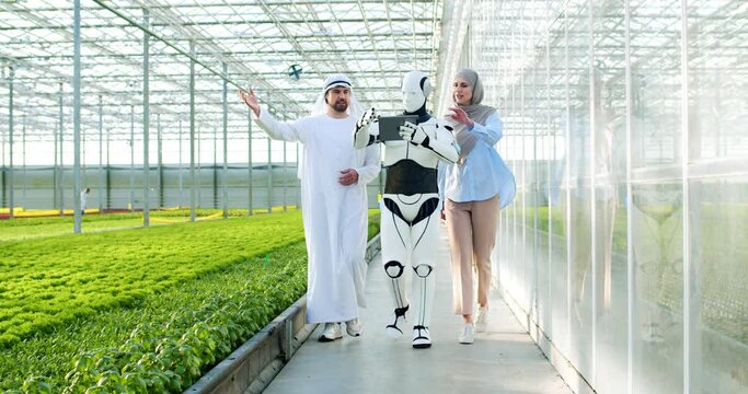 Muslim couple walking with human-like robot along polycarbonate wall of greenhouse. Cybernetic smart humanoid with digital tablet showing industrial glasshouse complex.