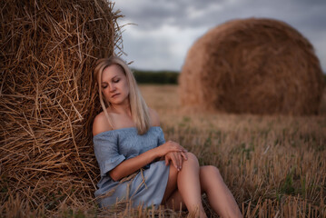 Portrait of a young beautiful blonde girl in rustic style resting on a haystack in a field. A young girl rests by a haystack in a field after the harvest.