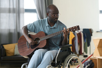 African American man in eyeglasses sitting in wheelchair and learning to play guitar during his leisure time at home