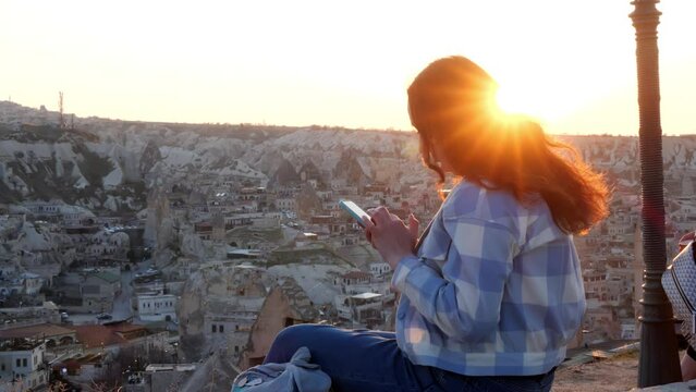 Rear view of woman tourist in blue and white check jacket sitting on top of mountain and typing text message or chatting. Sunbeam in hair. Top view of old town Goreme Cappadocia Turkey on sunset