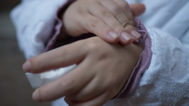 Close-up hands of little person applying moisturizer on palms. Unrecognizable Caucasian little woman in white bathrobe rubbing cream in skin