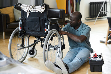 African man with disability sitting on floor and fixing his broken wheelchair with instruments