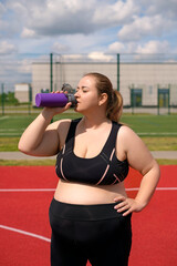 A fat plump woman in a black fitness tracksuit drinks water from a sports bottle quenching her thirst after working out on the street. Doing sports at an outdoor stadium on a summer morning