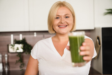 Beautiful plus size plump woman drinking healthy juice smoothie super food drink for vitamins at home kitchen, having breakfast looking at camera