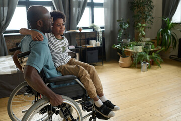 African disabled father riding his son on wheelchair while he sitting on his knees, they playing in...