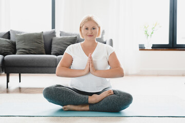 Fototapeta na wymiar Zen-like caucasian young plus size plump woman in fitness clothes meditating on sporty mat, sitting relaxing in yoga lotus position at home looking at camera