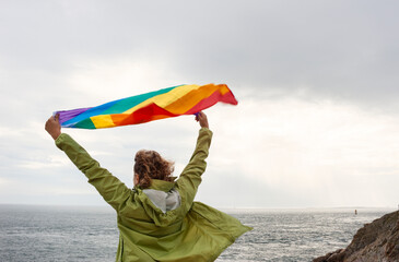 Unrecognizable blonde woman raising her arms at the tip of the cape waving a rainbow flag
