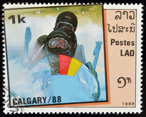 Cancelled postage stamp printed by Laos, that shows German Bob sleigh teram, promoting Winter Olympic games in Calgary, circa 1988. - obrazy, fototapety, plakaty