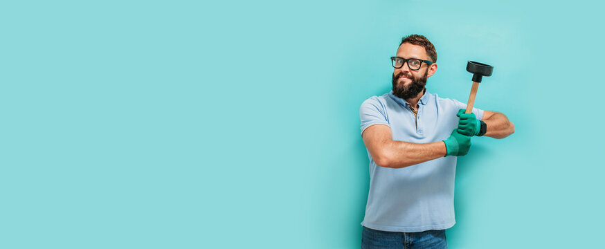 Young handsome man wearing plumber uniform holding toilet plunger looks happy. Professional cleaning of clogged pipes. Studio shot on blue background. Funny promotion poster