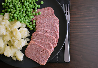 Sliced canned corned beef with boiled potatoes and peas on a black plate.