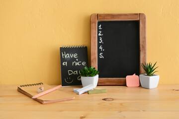 Chalkboard with empty to-do list, pen and notebook with text HAVE A NICE DAY on table near yellow...