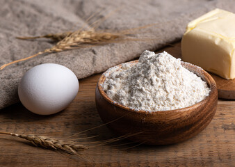 Flour in a wooden bowl, butter and an egg on a wooden table.