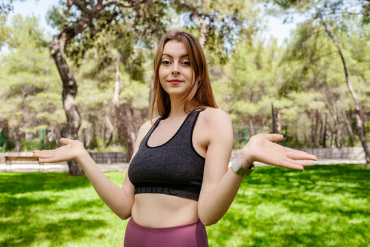 Brunette sportive woman wearing black sports bra standing on city park, outdoors clueless and confused with open arms, shrugging shoulders, no idea and doubtful face.