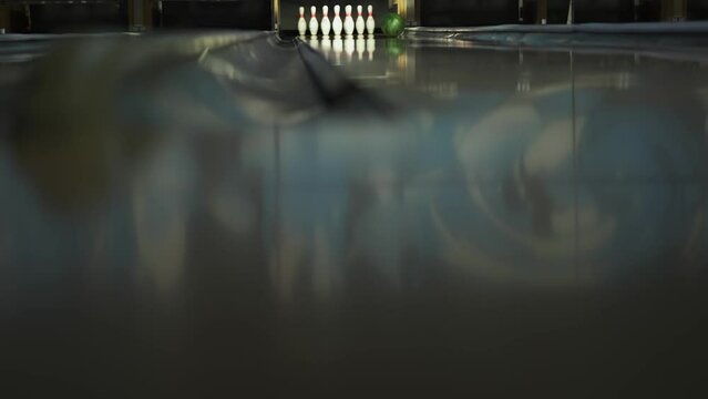 Green ball rolls down lane in the bowling alley. Ball knocks down half the pins in the pindeck, pinsetter swip bar descends. View from the track level, from the gutter.