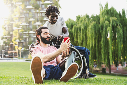 Two friends, one Hispanic with a prosthetic leg and one African in a wheelchair are relaxing outdoors chatting together and using smartphones and social networks - disability and integration concept