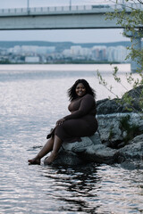 Vertical smiling playful barefoot curly plump full-figured Afro American interracial woman sit by the sea or river coast, wash legs and rest. Mixed race ethnicity, diversity. Windy summer weekends