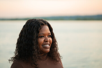 closeup portrait of curly african afro american dark skin woman looking away near lake outside. Interracial female on weekends. Mixed race people, ethnicity, diversity. Sunset horizon on background