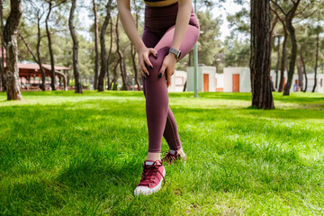 Brunette sportive woman wearing black sports bra standing on city park, outdoors touching injured knee with hands. Injured leg, healthy lifestyle and sport concept.