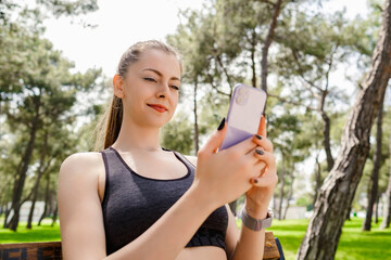 Cheerful brunette woman wearing sportive clothes on city park, outdoors looking at the phone screen and using phone. Messaging with friends, watching video or scrolling on social media.