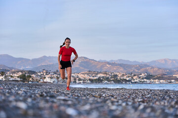 young athletic woman in a red shirt and braid running on the shore of the beach with mountains in...