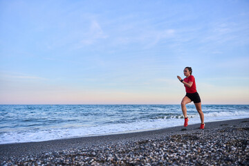 Fototapeta na wymiar young athletic woman in a red shirt and braid running on the shore of the beach with mountains in the background