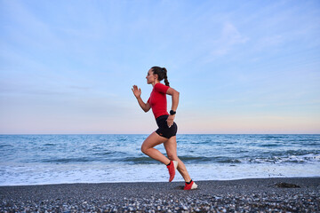 young athletic woman in a red shirt and braid running on the shore of the beach with mountains in the background