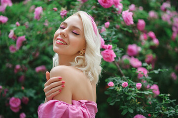 Fototapeta premium Outdoor portrait of happy smiling beautiful blonde woman wearing pink off shoulder blouse, headband posing in blooming rose garden. Copy, empty space for text 