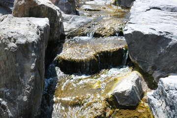 Fresh water creek flow with big boulder rocks and stones close up landscape photo. Rocky river shore natural background image with rough texture. Small waterfall stream with water glimmering in sun. 