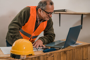 Construction contractor reviewing the development of the work on the laptop and the plans and lists on paper.