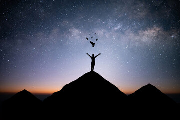 Young woman traveler standing alone on top of mountain and raise both arms praying and free bird enjoying nature on beautiful night sky, star, milky way background. Demonstrates hope and freedom.