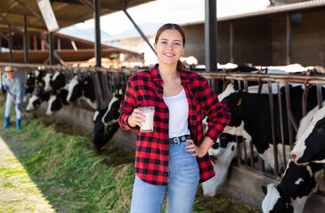 Fototapeta Portrait of cheerful young girl standing with glass of fresh milk near stalls with cows on dairy farm on sunny summer day obraz