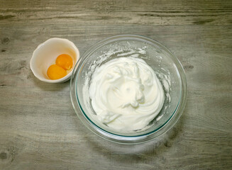 Two egg whites beaten in a glass bowl and the egg yolks on the side.