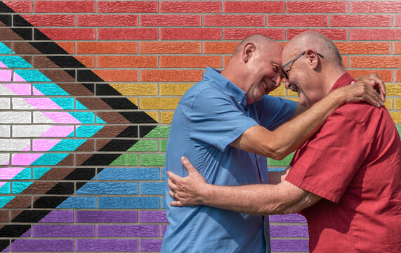 Mature Gay Couple Embrace In Front Of A Brick Wall Painted With The LGBT Progressive Pride Rainbow Flag