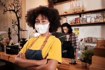 Portrait of young African American female barista with face mask looking at camera, arms crossed...