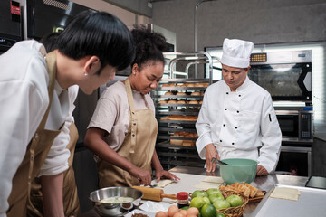 Cuisine course, senior male chef in cook uniform teaches young cooking class students to knead and...